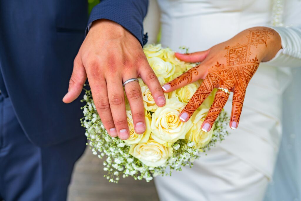 Close-up of a couple's hands with wedding rings and henna designs, holding a bouquet.